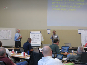 Two men present information on a large screen, in front of a group.