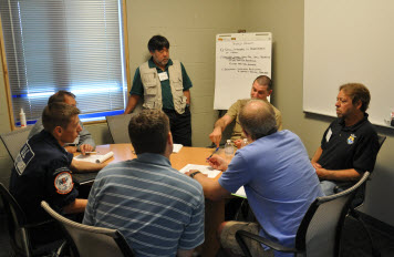 A group of men engaged in discussion around a table, next to a poster board.